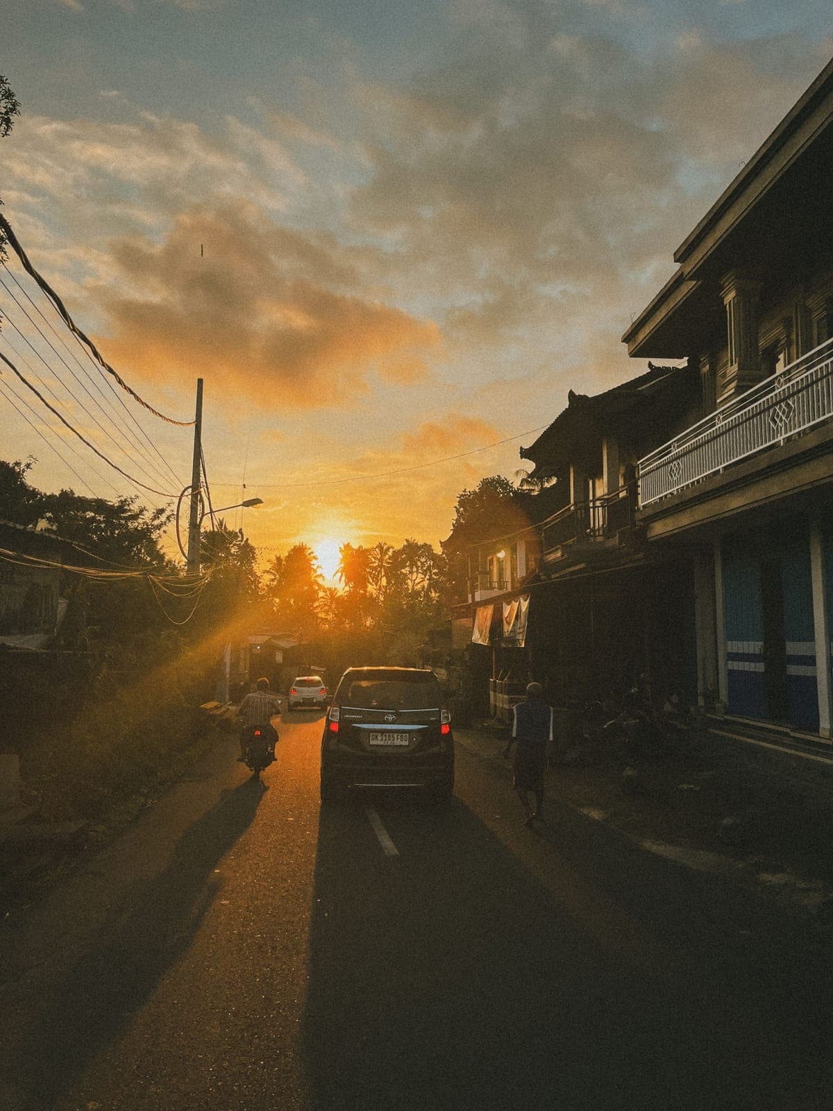 Village street backlit by the setting sun, with a car and a scooter